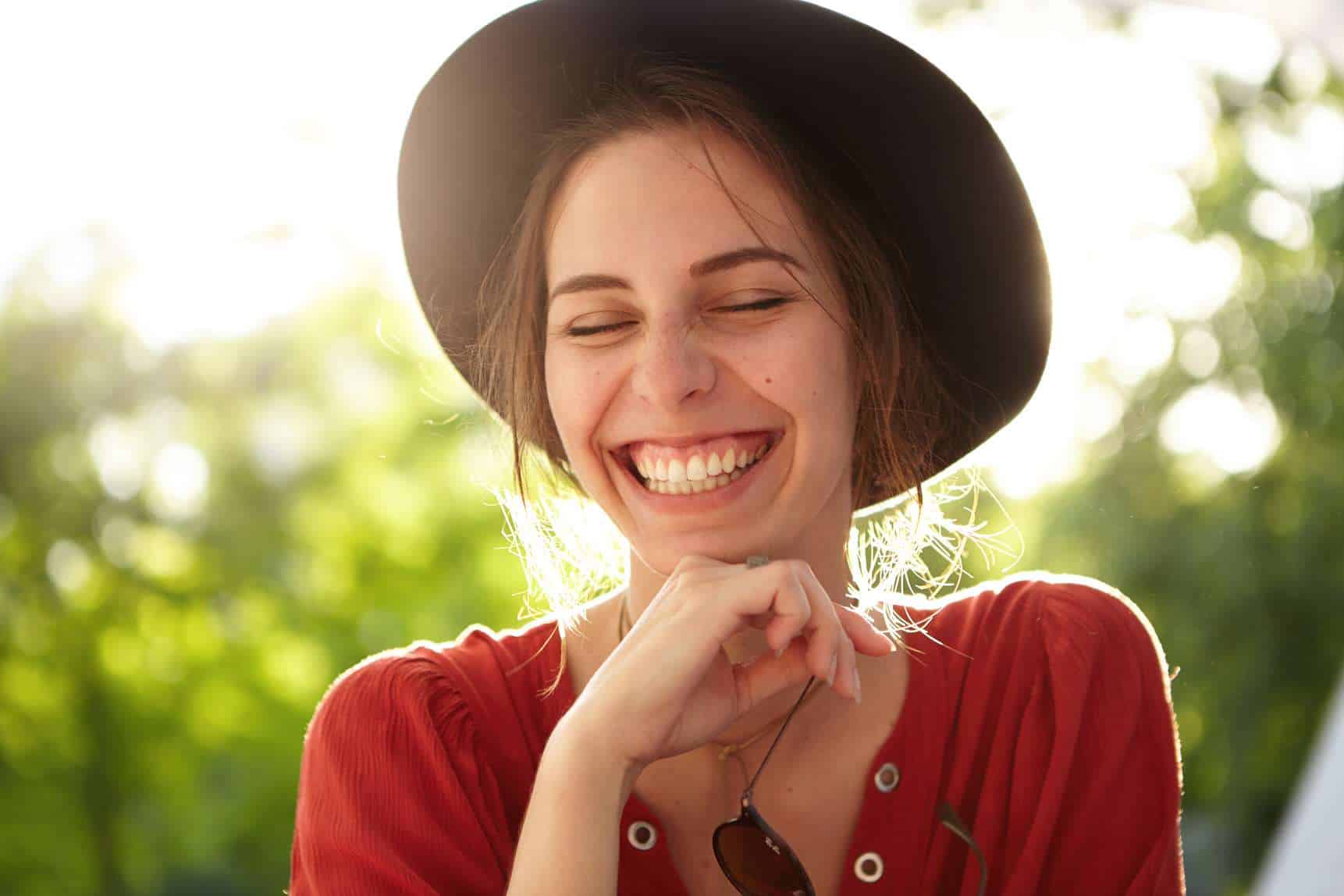 Close-up of gentle woman wearing big black summer hat and casual red shirt closing her eyes with pleasure smiling with joy being happy to spend nice warm summer day in park breathing fresh air