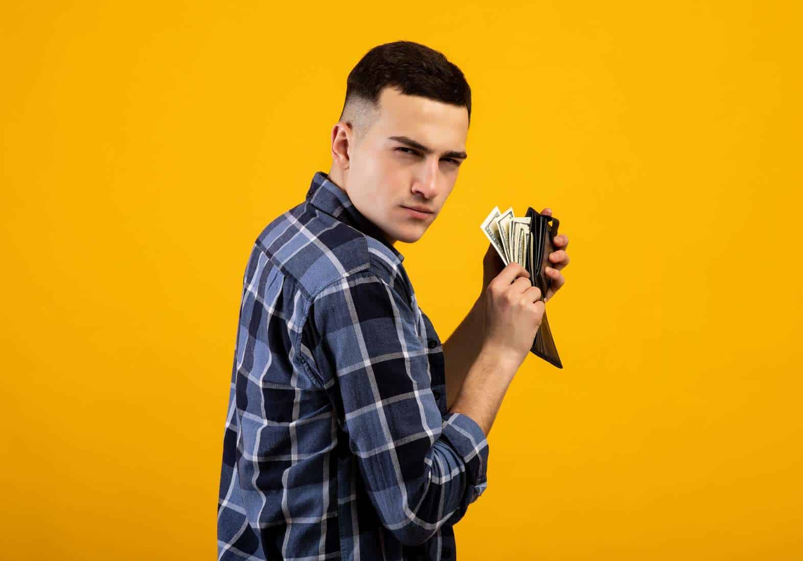 Young man with wallet full of money being greedy over his wealth on orange studio background